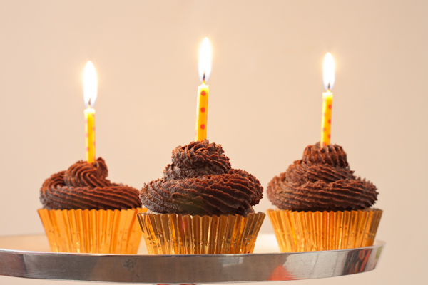Three vanilla cupcakes with lit birthday candles set in fudgy frosting swirls.